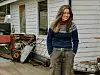 Kvinne står foran et hus med trillebår, verktøy og ved langs ytterveggen.