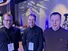 Tre personer med konferansebadge står i en blåbelyst konferansesal.