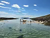 Sommerdag ved grunt, klart kystvann med mennesker og kajakk på sandbanke mellom svaberg.