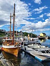 Tre- og motorbåter fortøyd ved brygge i småbåthavn med blå himmel og skyer.