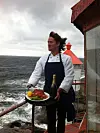 Servitør holder et brett med mat og vin ved en kyst med røft hav og grå himmel.