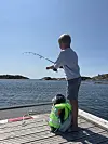 Et barn fisker fra en brygge med en hund i grønn vest ved siden av, mot sjø og klar himmel.