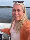 Kvinne i oransje cardigan står på en båt med sjø og kystlinje bak seg.