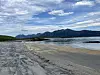 Grå sandstrand ved lavvann med fjell og skyet himmel i bakgrunnen.