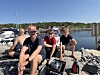 Fire personer sitter på svaberg ved en småbåthavn med båter og klart vann i bakgrunnen.