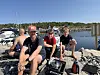 Fire personer sitter på svaberg ved en småbåthavn med båter og klart vann i bakgrunnen.