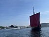 Tradisjonell båt med rødt seil på vannet, med en passasjerferge i bakgrunnen.