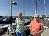 To personer står ved en marina med seilbåter og master i solskinn.