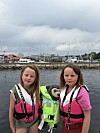 To barn i redningsvester står ved en havn med en bamse i en grønn vest mellom seg.
