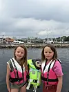 To barn i redningsvester står ved en havn med en bamse i en grønn vest mellom seg.