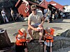 Mann sitter på en stein med to barn i redningsvester foran en rød bygning ved havnen.