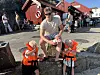 Mann sitter på en stein med to barn i redningsvester foran en rød bygning ved havnen.