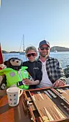 Par med teddybjørn ved et bord på en marina, med seilbåter og åser i bakgrunnen.