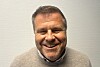 Nærbilde av en smilende mann i grå genser foran en lys vegg.